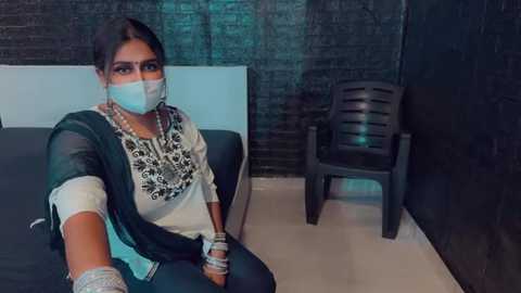 Media: Video of a South Asian woman with medium skin tone, wearing a white face mask, a white blouse, and a green shawl, sitting on a bed with a dark headboard, in a dimly lit room with a black brick wall and a black plastic chair.