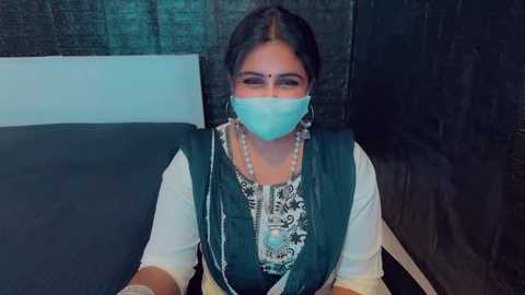 Media: Video of a South Asian woman with medium skin tone, wearing a white saree, a blue face mask, and silver jewelry, seated on a dark green couch against a black textured wall.