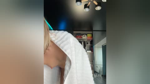 Media: A video of a blonde woman in a white towel, partially hidden, standing in a dimly lit, cluttered room with a black ceiling, white shelves, and a fan.