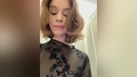 Media: Video of a young woman with shoulder-length, wavy, light brown hair, wearing a sheer black blouse with floral patterns, standing indoors in a blurred background.