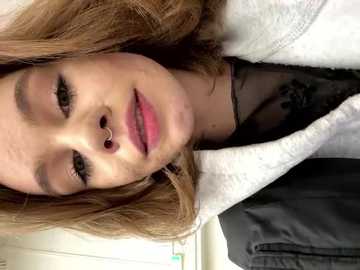 Media: A close-up video of a young woman with fair skin, long brown hair, and full lips, wearing a white sweater and black top, with a nose ring, taken indoors.