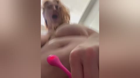 Media: A blurry video of a nude, overweight woman with large breasts and light skin, using a pink vibrator on her left nipple. She has wavy, shoulder-length blonde hair and is in a bedroom setting.