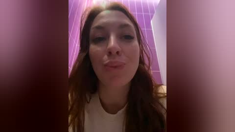 Media: Video of a young woman with long, wavy brown hair, making a pouty face against a purple and white tiled bathroom background.