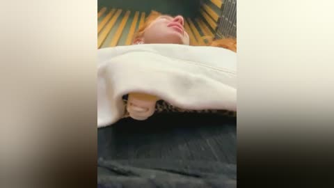 Media: Video of a baby with pale skin and red hair, covered in a white blanket, lying on a wooden floor with a yellow pattern.