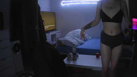 Media: Video of a dimly lit bedroom with a woman in black lingerie adjusting a flat-screen TV on a dresser. A bed with a rumpled blue comforter is visible, and a desk with a laptop and drinks is in the foreground.