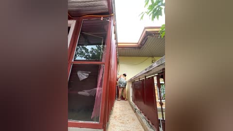 Media: Video of a narrow, red-painted outdoor structure with a glass door, reflecting greenery. Two people in casual clothes stand in front, next to a beige building with a tiled roof.