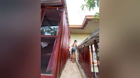 Media: Video of a woman with long hair, wearing a tank top and shorts, standing on a narrow, red-painted balcony railing, overlooking a tiled patio.