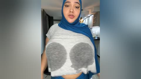 Media: Video of a woman with large, exaggerated breasts, wearing a blue hijab and gray crop top with large gray circles, in a modern kitchen.