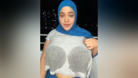 Media: Video of a young woman with medium skin tone, wearing a blue hijab and a gray crop top with a large, visible nipple, taken outdoors at night with a cityscape background.