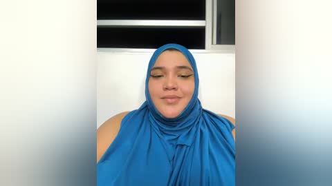 Media: Video of a smiling woman with light skin, wearing a bright blue hijab, indoors against a white wall with a window.