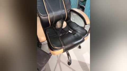 Media: Video of a black leather office chair with tan stitching and a large beige cushion in the seat. The chair is in an office setting with light-colored flooring and a blue curtain in the background.