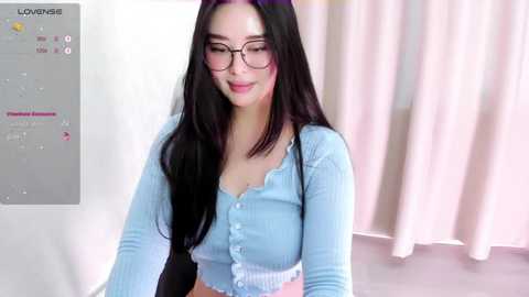 Media: Video of a smiling Asian woman with long black hair, wearing glasses and a light blue ribbed cardigan, standing in a room with white curtains.