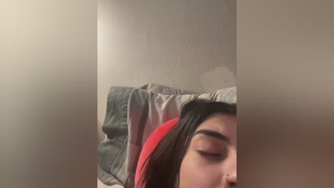 Media: Video of a young woman with closed eyes, lying on a bed with a red pillow and white sheets, against a beige wall with a textured finish.