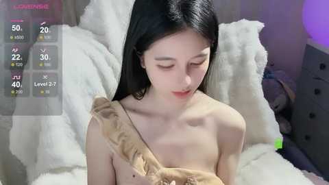 Media: A video of a fair-skinned, petite East Asian woman with long black hair, wearing a beige off-shoulder dress, sitting on a white fur blanket. The background features a gray dresser and a plush toy.