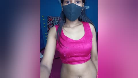 Media: A video of a young woman with medium brown skin, wearing a pink crop top, black face mask, and black hair. She stands against a blue wall with patterned fabric in the background.