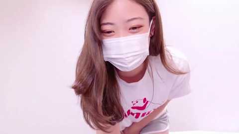 Media: Video of a young Asian woman with long brown hair, wearing a white mask, white T-shirt with a graphic, and gray shorts, leaning forward with hands clasped.