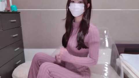 Media: Video of an East Asian woman with long black hair, wearing a pink face mask and matching long-sleeve top and pants, sitting on a white toilet in a modern bathroom.