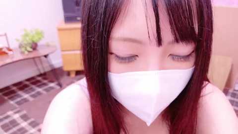 Media: A video of an Asian woman with long, straight, dark hair and bangs, wearing a white surgical mask, sitting indoors on a plaid carpet.