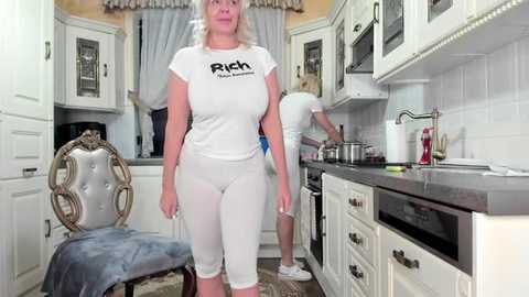 Media: Video of a busty, blonde woman in a tight white tank top and leggings, standing in a chic, white kitchen with stainless steel appliances.