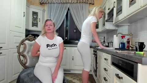 Media: Video of a white woman in a tight white T-shirt, seated in a kitchen with beige walls, next to a blonde woman in white pants, leaning against a counter.