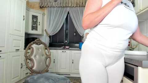 Media: Video of a curvy woman in a tight white bodysuit, standing in a modern kitchen with white cabinets, a silver chair, and a window with sheer curtains.