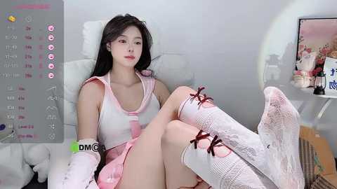 Media: Video of an Asian woman with long black hair, wearing a pink sailor outfit, sitting on a bed, with a digital clock and a white teddy bear visible in the background.