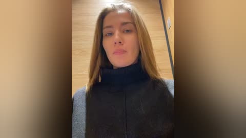 Media: Video of a young Caucasian woman with long blonde hair, fair skin, and a neutral expression, wearing a black turtleneck sweater, standing in a beige hallway with wooden floors.