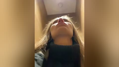 Media: A video of a young girl with long brown hair, wearing a dark sweater, looking upwards with a serene expression, inside a dimly lit, narrow hallway with beige walls and a recessed ceiling light.