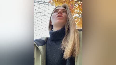 Media: Video of a young Caucasian woman with long, blonde hair, wearing a dark turtleneck sweater and green jacket, standing outdoors under a wire fence with autumn leaves in the background.