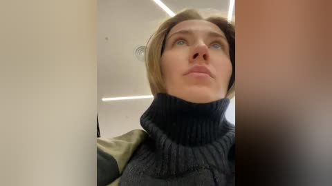 Media: Video of a woman with short blonde hair and blue eyes, wearing a dark gray turtleneck sweater, standing in a dimly lit, narrow hallway with beige walls.