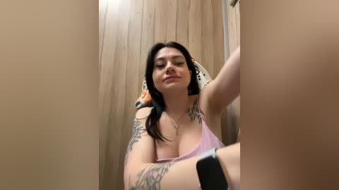 Media: A video of a young, light-skinned woman with long black hair, tattoos on her arms, and a pink tank top, sitting in a wooden stall, taking a selfie.