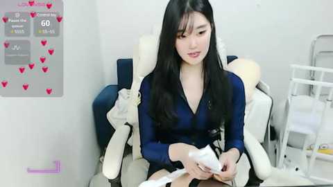 Media: Video of a young Asian woman with long black hair, wearing a blue V-neck shirt, applying makeup to a man reclining in a white massage chair. The background includes a wall chart with heart icons and a white medical cabinet.