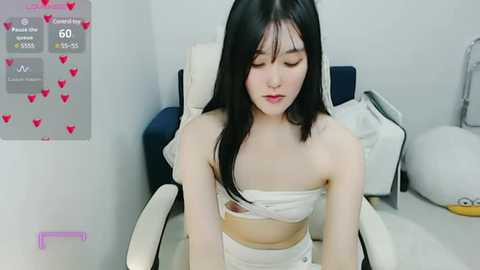 Media: A video of a young Asian woman with straight black hair, wearing a white strapless top revealing her midriff, sitting in a white chair in a minimalistic room.