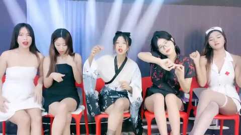 Media: Video of five Asian women in formal dresses, white, black, and floral, sitting on red chairs, hands on faces, looking distressed. Background is a blue and white curtain.