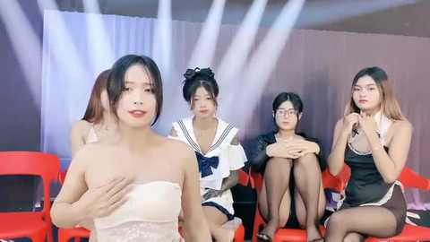 Media: Video of five Asian women in revealing outfits, sitting on red chairs, holding microphones, with stage lights in the background.