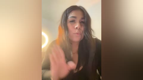 Media: Video of a young woman with medium brown skin, long dark hair, and a neutral expression, reaching out with her hand. She wears a black top. Background is blurred with warm, ambient lighting.