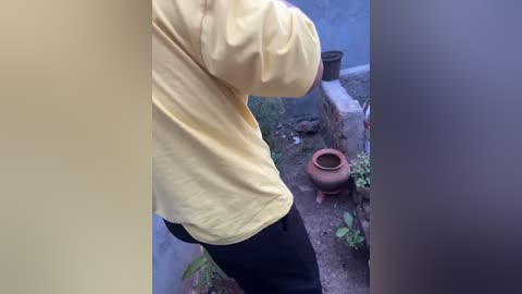 Media: Video of a person in a yellow shirt and black pants standing near a red pot and green plants on a dirt ground, beside a stone wall, with a blurred background.