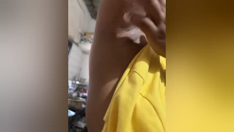 Media: A close-up video of a person's upper torso, partially covered by a yellow garment. The background features a cluttered, dimly lit room with shelves and various objects. The person's skin tone is light, and their hand is visible holding the garment.