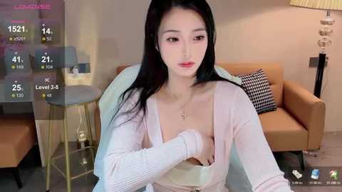 Media: Video of an East Asian woman with long black hair, wearing a low-cut pink cardigan, seated in a modern living room.