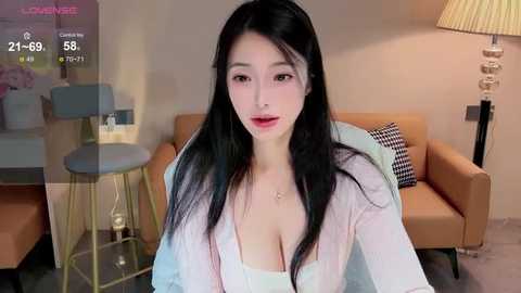 Media: A video of an Asian woman with long black hair, wearing a low-cut pink shirt, sitting in a modern living room with beige furniture.