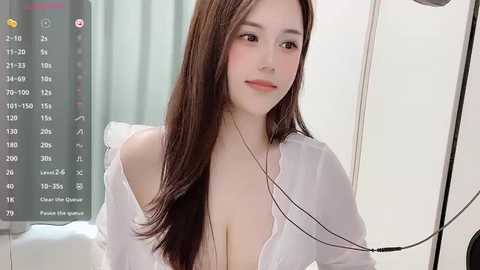 Media: Video of an East Asian woman with long, straight brown hair and fair skin, wearing a white, off-shoulder blouse, sitting in a hospital room with a medical monitor and IV pole in the background.