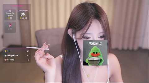 Media: Video of an Asian woman with long brown hair, holding chopsticks, wearing headphones, overlaid with a digital green alien character, in a beige room.