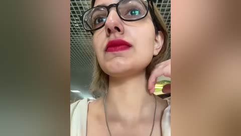 Media: Video of a woman with light skin, wearing glasses, red lipstick, and a beige top, against a grid ceiling background.