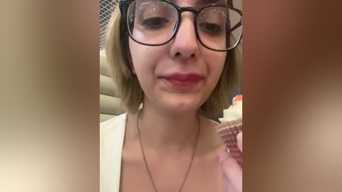 Media: A close-up video of a young woman with fair skin, light brown hair, and large black-framed glasses, holding a cupcake. She has a neutral expression, wearing a white top. Background is blurred.