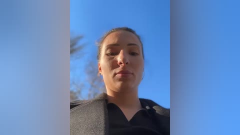 Media: Video of a woman with fair skin and brown hair in a bun, wearing a black shirt, looking directly at the camera against a bright blue sky.