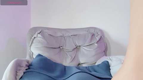 Media: A video of a woman's backside, lying on a bed with a rumpled, light grey pillow and a blue blanket partially covering her. The background is a plain white wall.