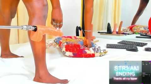 Media: Video of a nude, dark-skinned man inserting a realistic, flesh-toned dildo into a white bed. Background includes a colorful blanket, black sex toys, and a computer screen displaying \"STREAM ENDING\" text.