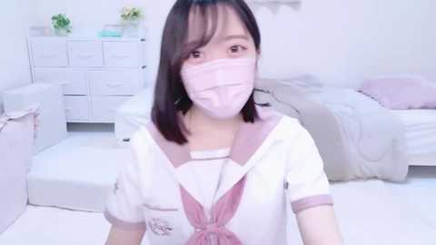 Media: A video of an East Asian woman with light skin and shoulder-length black hair, wearing a pink face mask, white sailor uniform, and pink bow, in a minimalist, white bedroom with two beds and potted plants.