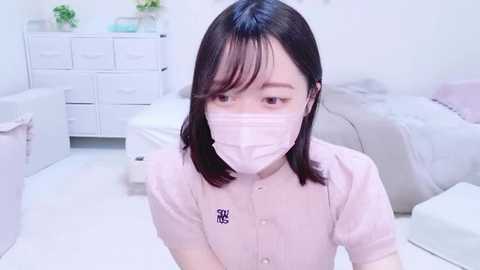 Media: Video of an Asian woman with shoulder-length black hair, wearing a pink medical mask, seated in a white, minimalistic room with white furniture, including a bed and drawers.