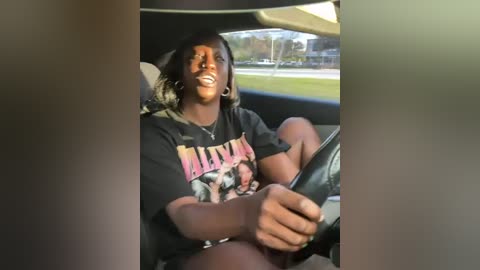 Media: Video of a joyful, dark-skinned woman with a medium build and shoulder-length, wavy hair, wearing a black graphic t-shirt and hoop earrings, driving a car.